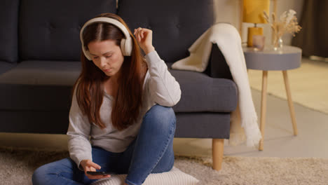 Mujer-Con-Auriculares-Inalámbricos-Sentada-En-El-Suelo-En-Casa-Transmitiendo-Música-Desde-Un-Teléfono-Móvil-12